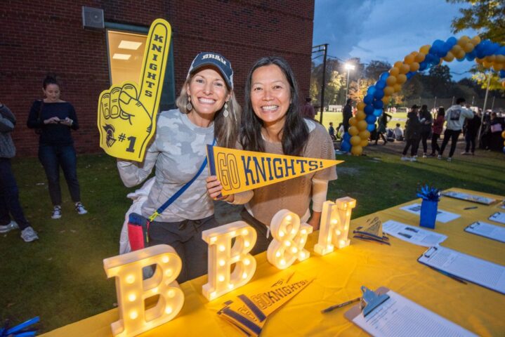 Two smiling women stand behind a table decorated with illuminated "B&N" letters and school spirit items, including a foam finger and pennant, at an outdoor event with people in the background.