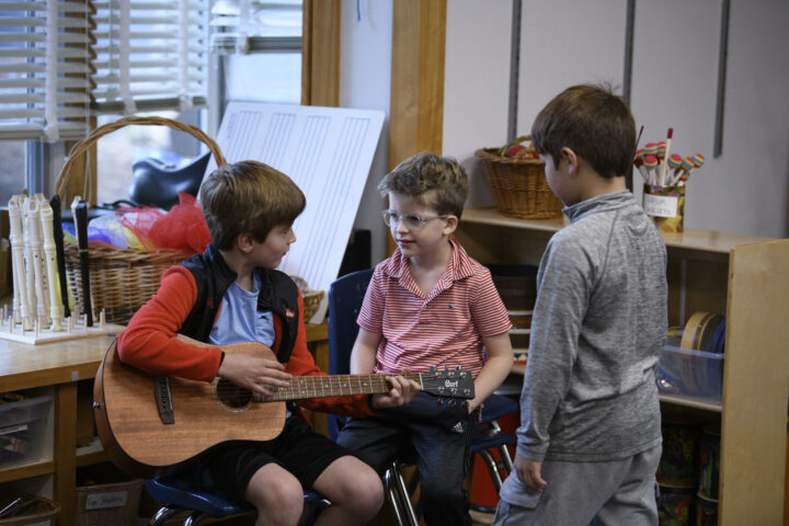 Three young boys are gathered in a room; one is seated, playing an acoustic guitar, and the other two are standing, watching him.