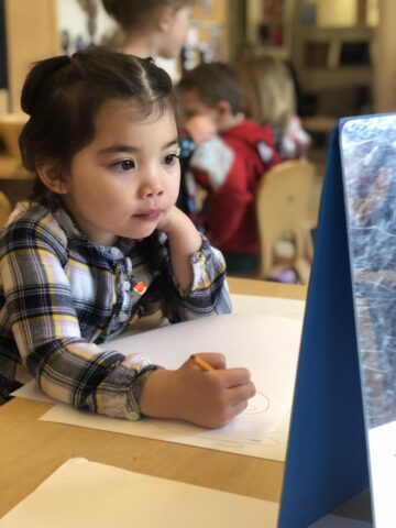 A young child sits at a table, looking at a blue easel while holding a pencil and resting her head on one hand.