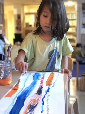 A child with long hair concentrates while painting colorful lines and patterns on a large sheet of paper, using a paintbrush.