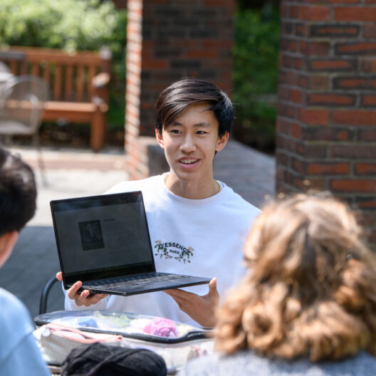 student showing laptop to other students outside.