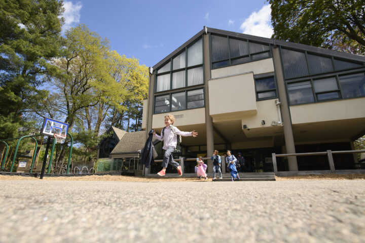 Children running and playing in front of a modern building with a basketball hoop and trees in the background.
