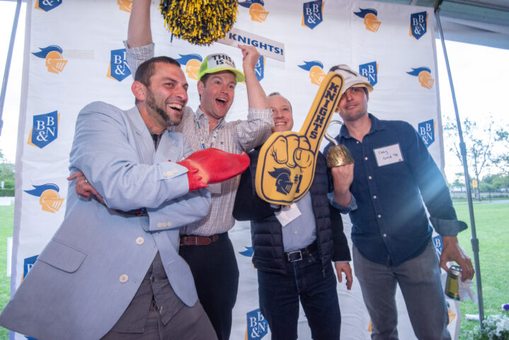 Four men wearing hats pose enthusiastically with Knights-themed props including a foam finger and pom-poms in front of a BB&N banner.