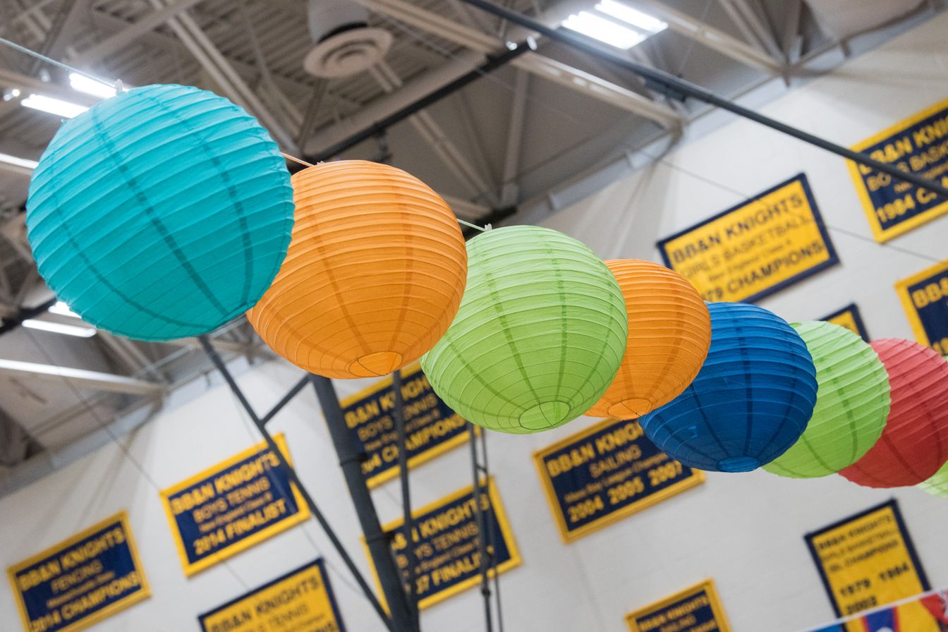 Seven colorful paper lanterns hang in a line in a gymnasium, with banners on the wall in the background.