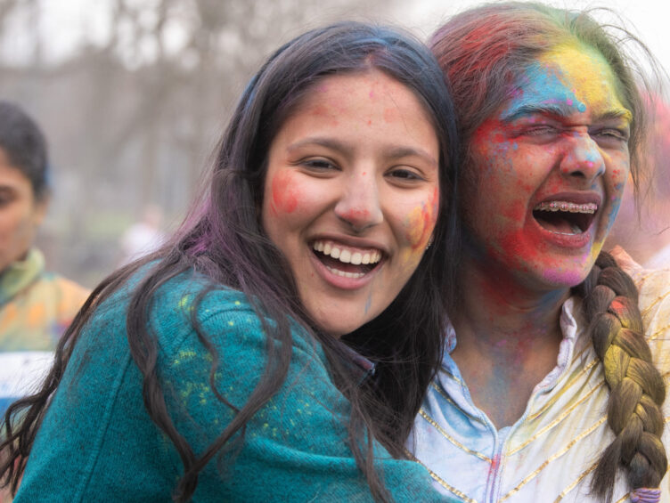 Two people covered in colorful powder smile and embrace outdoors during a celebration.