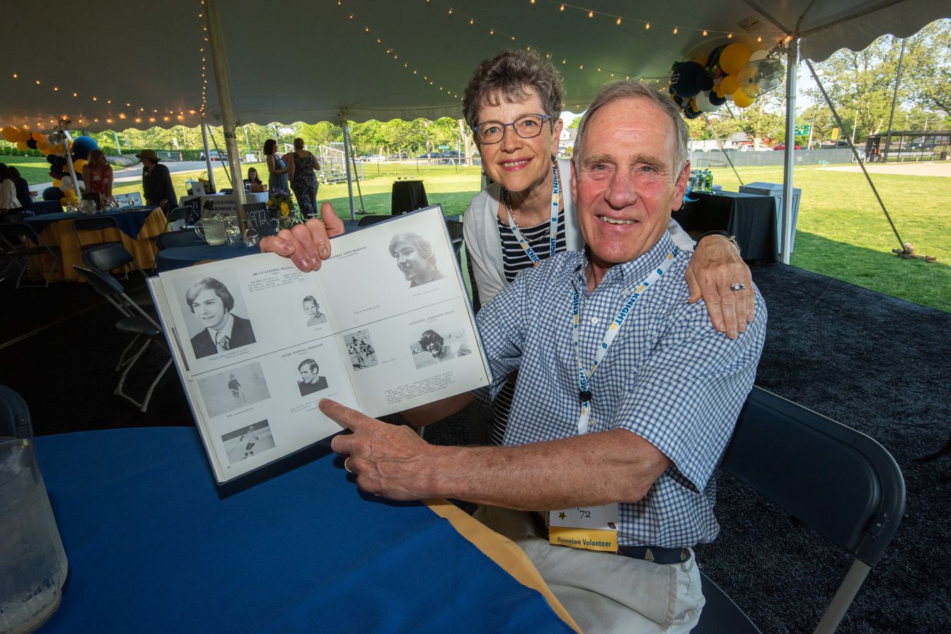 A man and woman smile as the man points to photos in a yearbook. They are seated at a table under a tent at an outdoor event, with decorations and other attendees in the background.