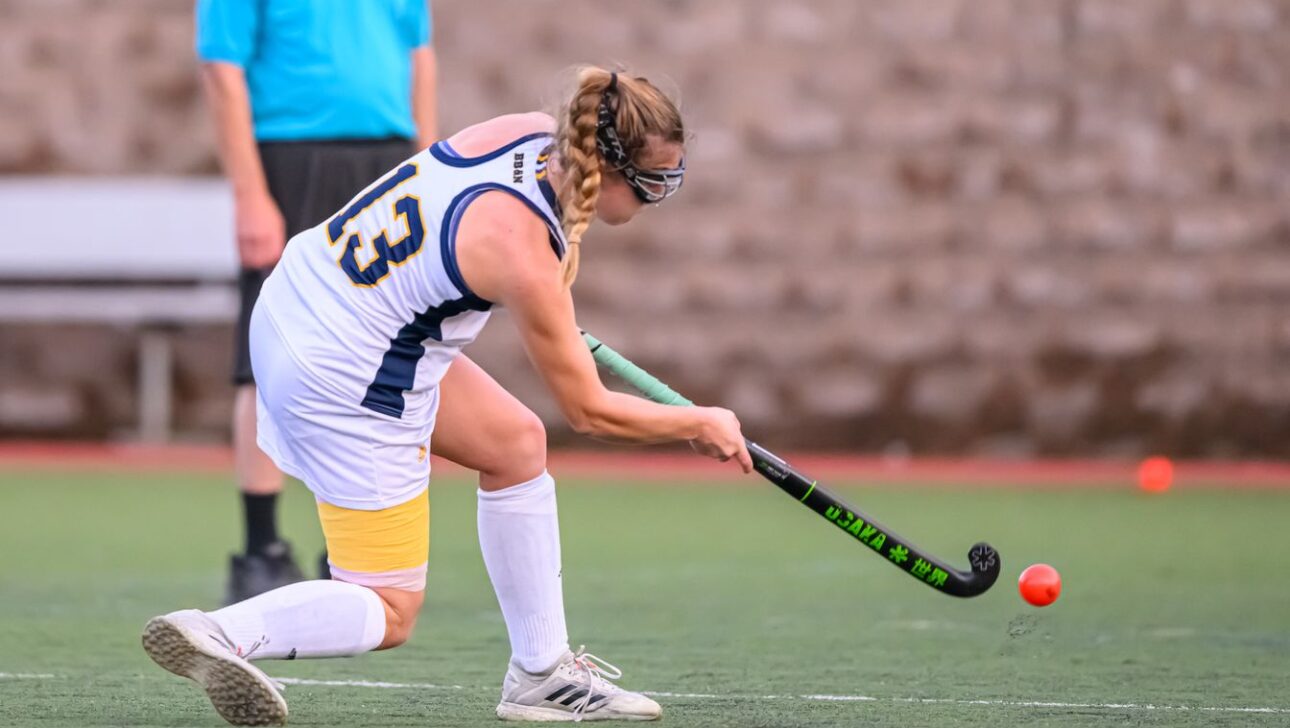A field hockey player wearing a white and blue uniform strikes a red ball on a grass field during a game.