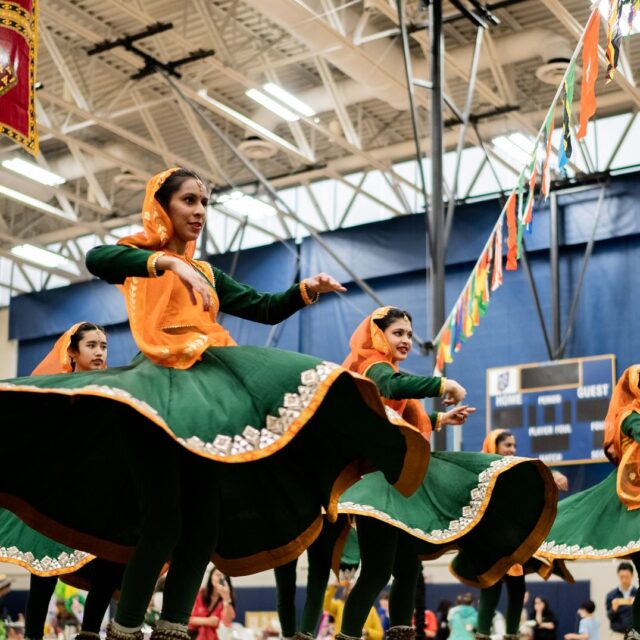 Dancers in green and orange traditional attire perform a folk dance in a decorated indoor venue with colorful banners overhead.