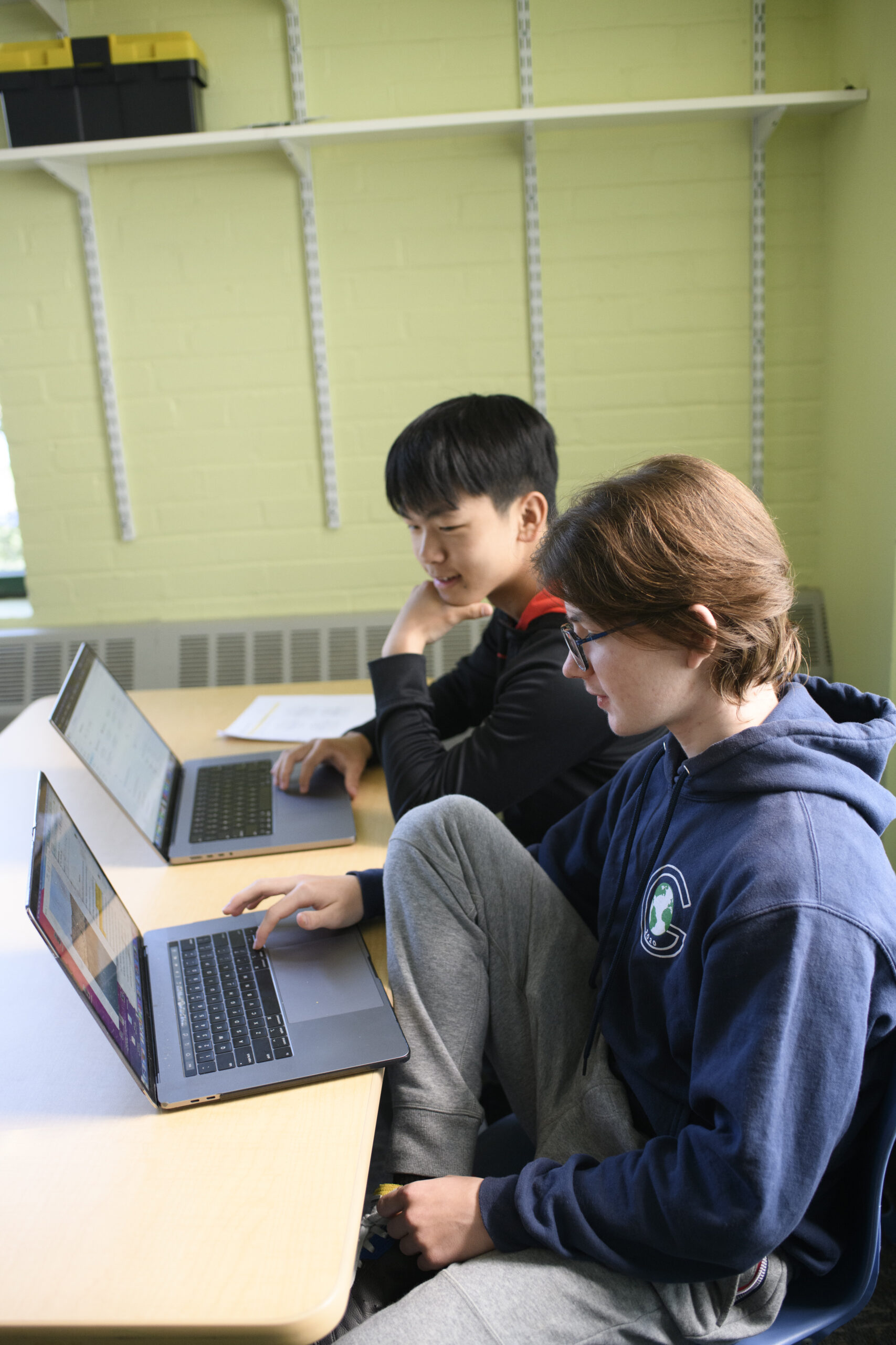 Two students sit at a table, each working on a laptop.