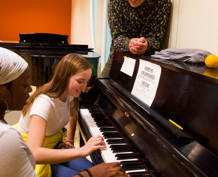 Three people are gathered around a piano; one is playing while the others watch.