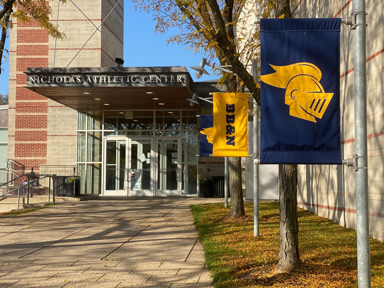 Entrance of Nicholas Athletic Center with glass doors, surrounded by light-colored brick walls and blue and yellow flags featuring a knight's helmet design and the initials "BB&N".