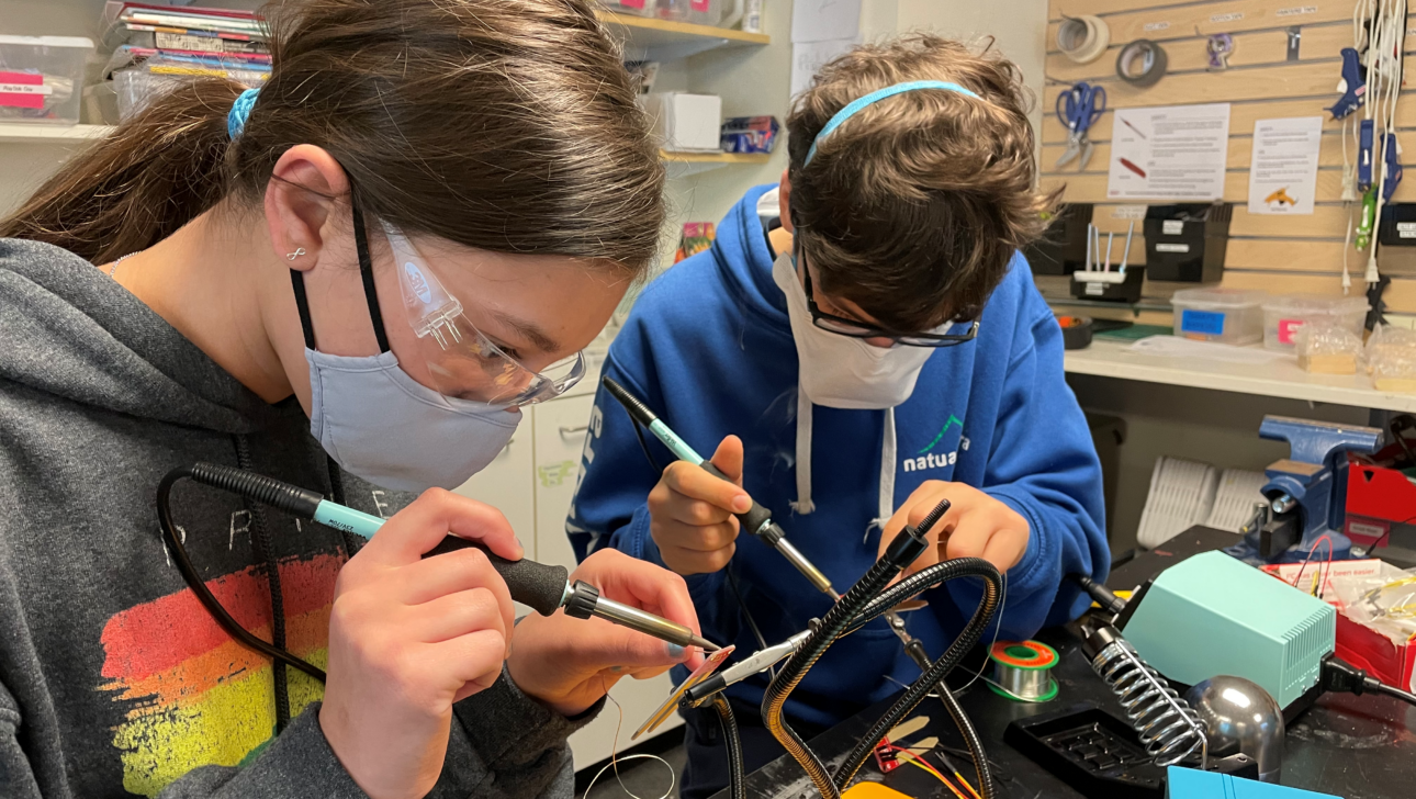 Two people focused on using soldering irons at a workbench, wearing safety glasses and face masks.