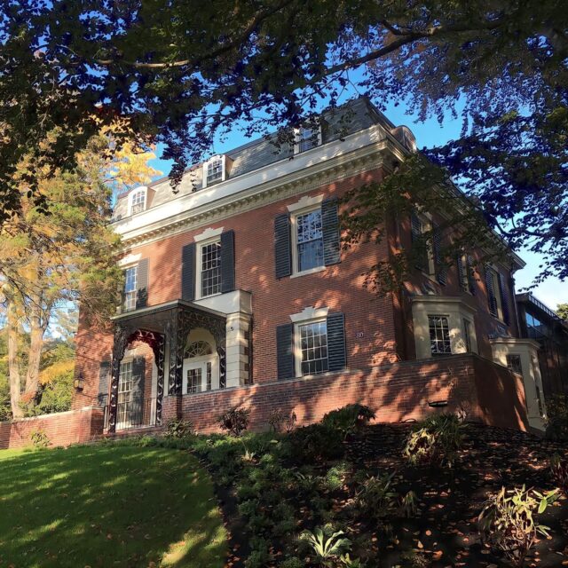 A two-story brick house with white trim, dark shutters, and a steep roof stands amidst tall trees and a well-maintained lawn on a sunny day.