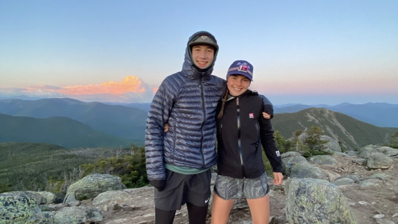 Two people in outdoor clothing standing on a rocky mountain top with a scenic view of mountains and a colorful sky in the background at sunset.