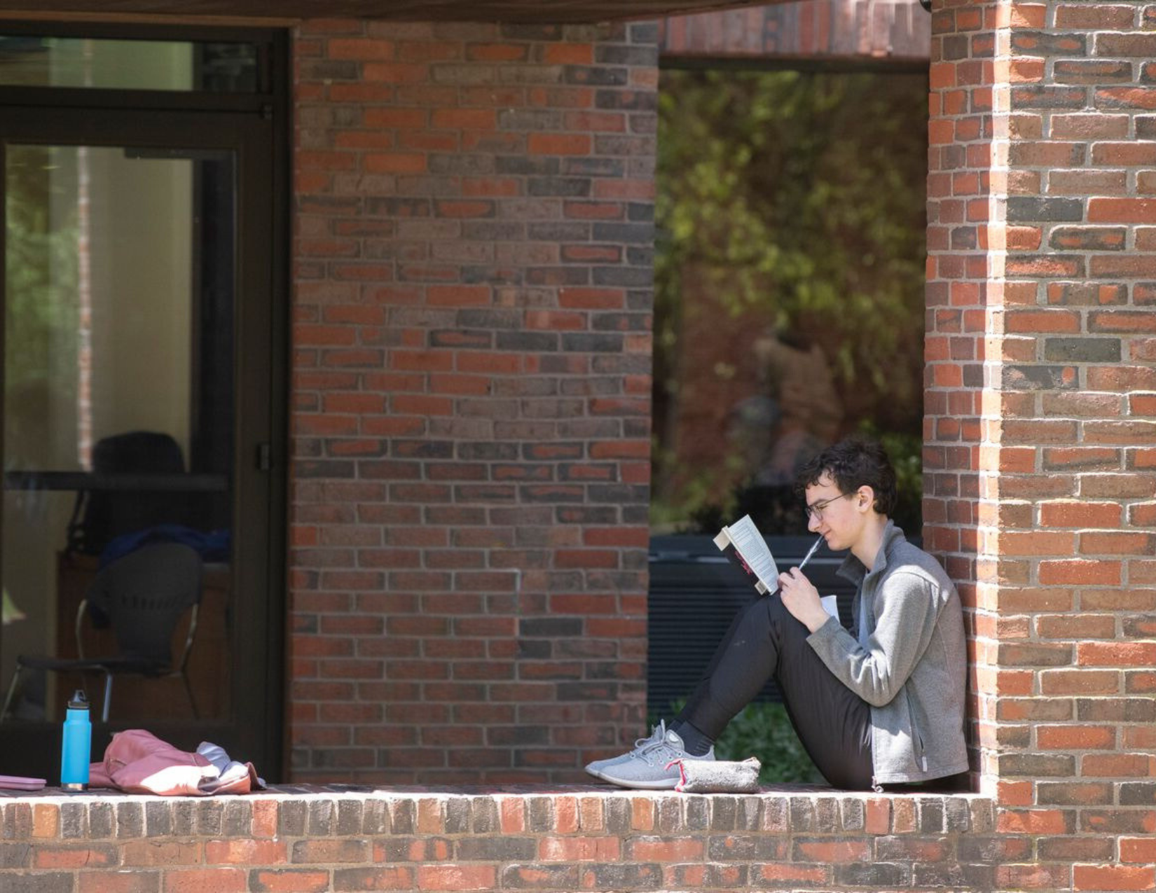 A student sits on the ledge of a brick building reading a book.
