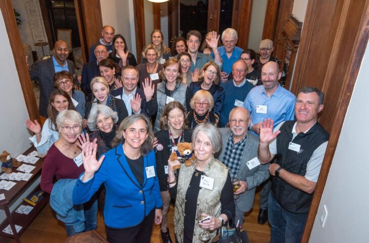 A group of people standing indoors, smiling and waving at the camera.