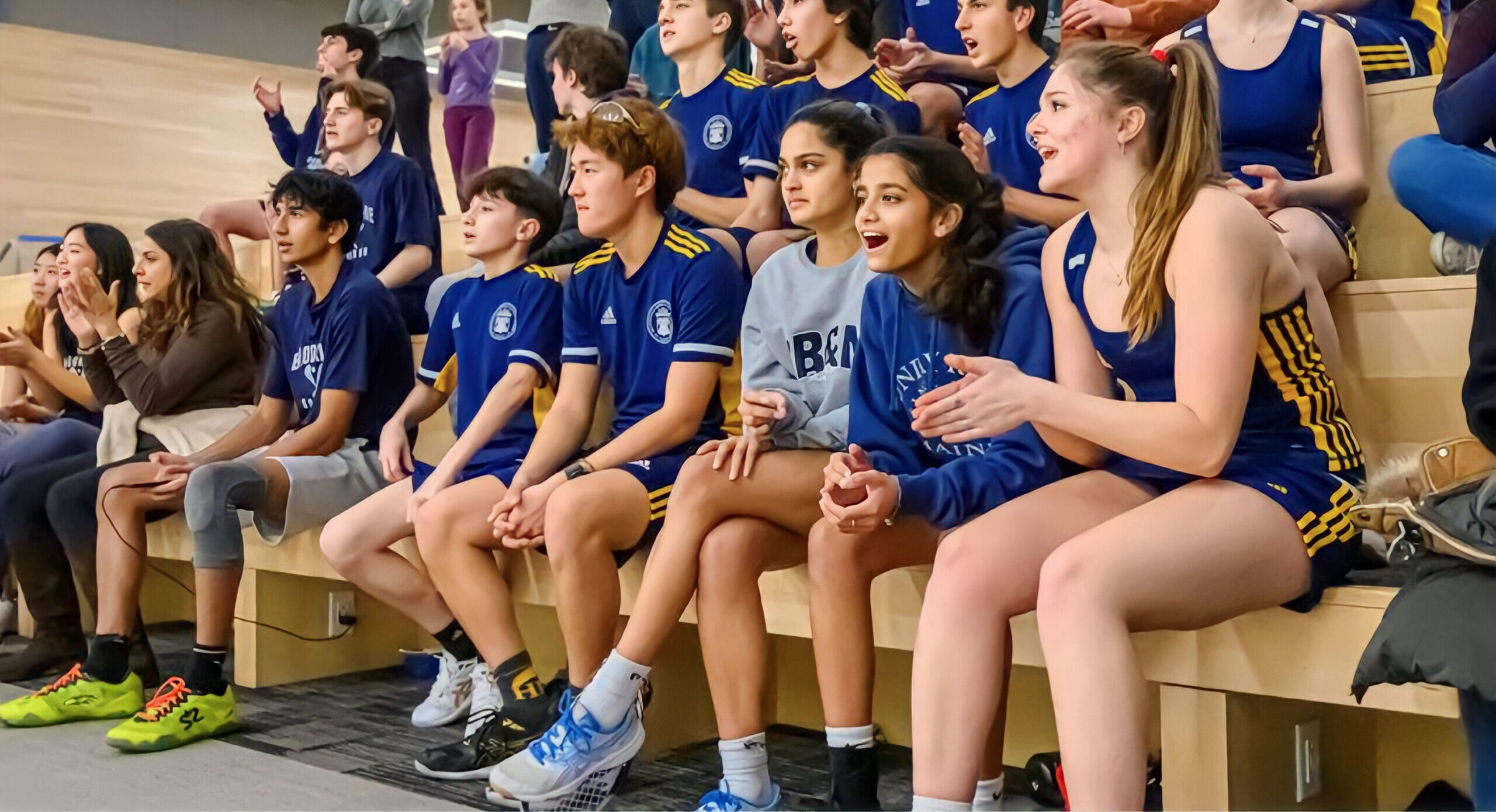 A group of students in sports uniforms sit on bleachers, clapping and cheering, while others around them watch a live event.