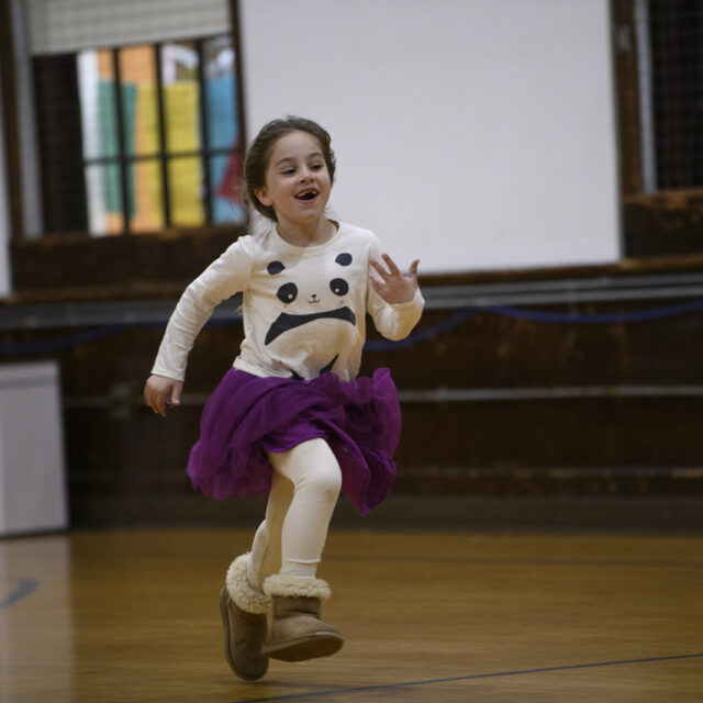 A child wearing a panda shirt, purple tutu, and boots runs indoors, smiling and looking to the side.
