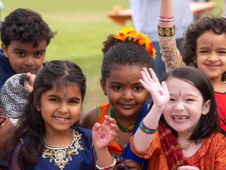 A group of children dressed in colorful traditional attire, some with bindi on their foreheads, smile and wave at the camera.