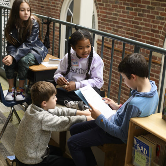 Four children are sitting and standing around a shelf, looking at a piece of paper held by one of the boys.