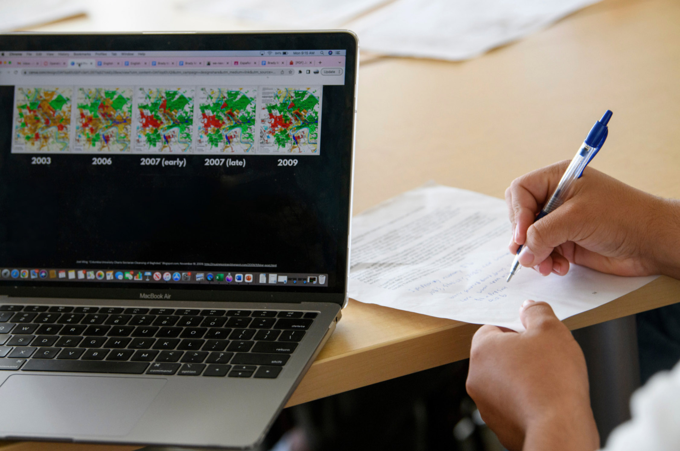 A person writes notes on paper while a laptop screen displays a series of maps labeled with different years from 2003 to 2009.