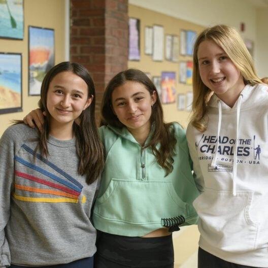 Three smiling girls stand together in a school hallway, with colorful artwork displayed on the wall behind them.