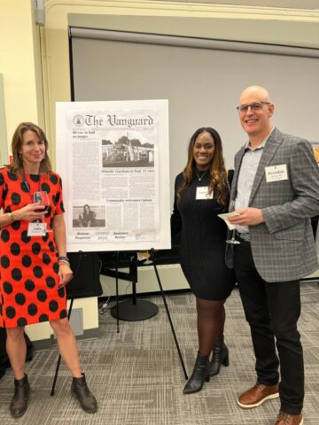 Three people are standing and smiling in front of an easel displaying a large newspaper front page.