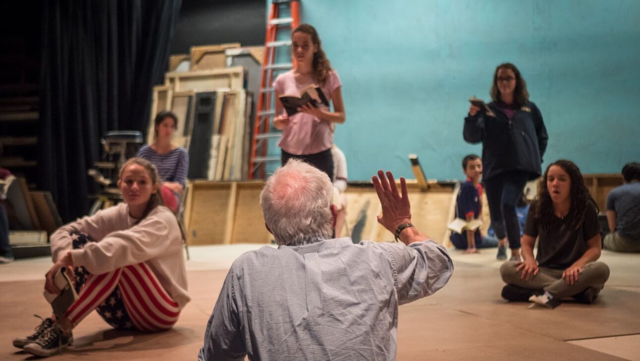 A person with white hair gestures while speaking to a group of people seated on the ground in what looks like a rehearsal space.