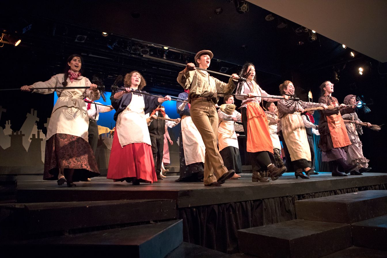 A group of performers on stage dressed in period costumes sings and dances energetically while holding onto a long metal bar.