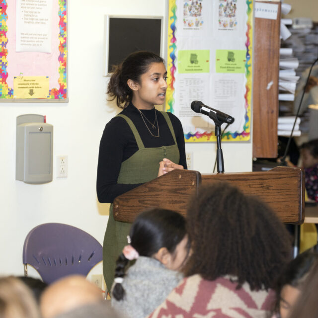 A woman speaks at a podium in a classroom. Students sitting in chairs listen while colorful posters and stacks of papers are visible in the background.