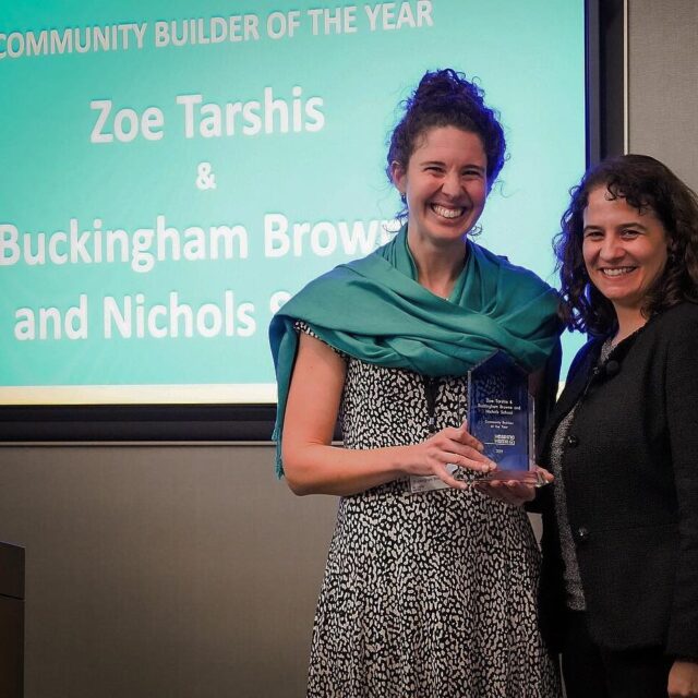Two women standing in front of a screen displaying "Community Builder of the Year," holding an award, smiling. The text on screen includes "Zoe Tarshis" and "Buckingham Brown and Nichols School.