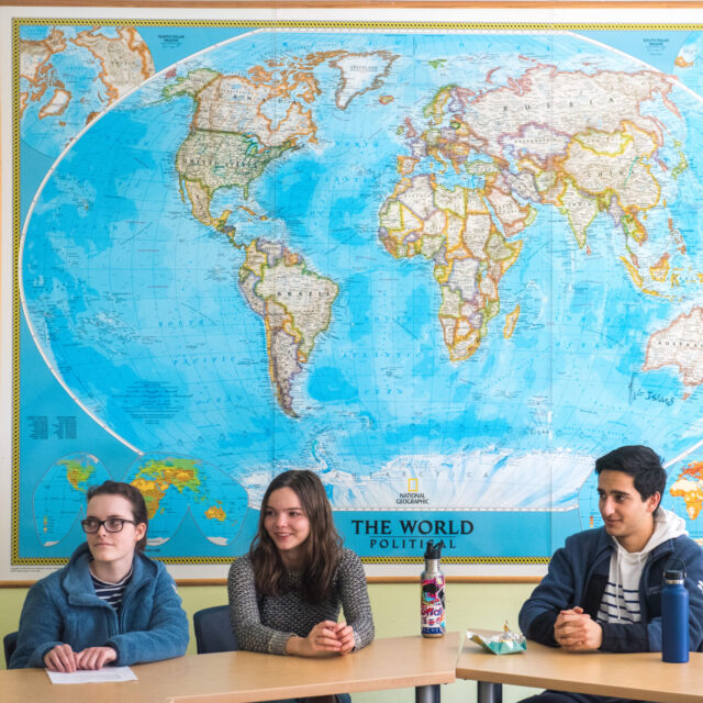 Three students sit at a table with notebooks and water bottles in front of a large wall map of the world in a classroom setting.