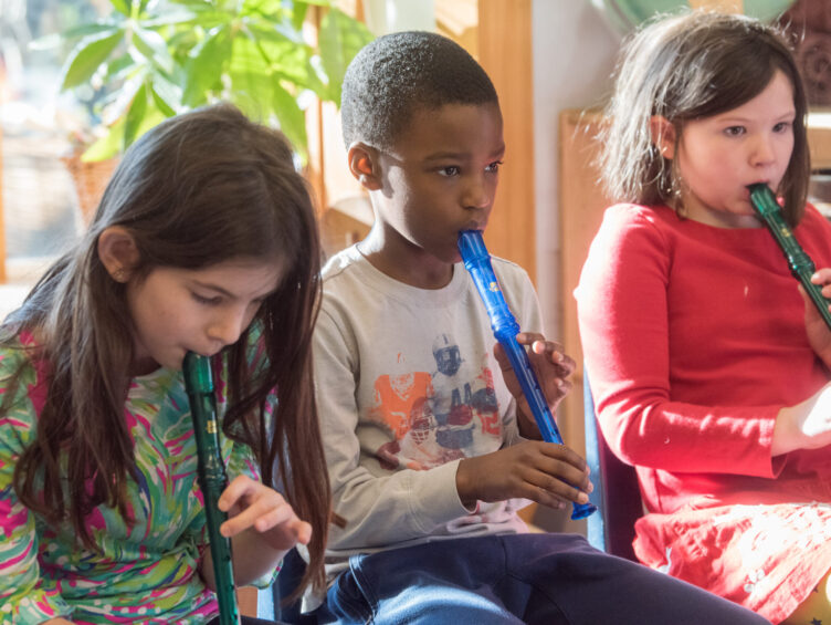 Three children sit in a row playing blue plastic recorders in a classroom setting.