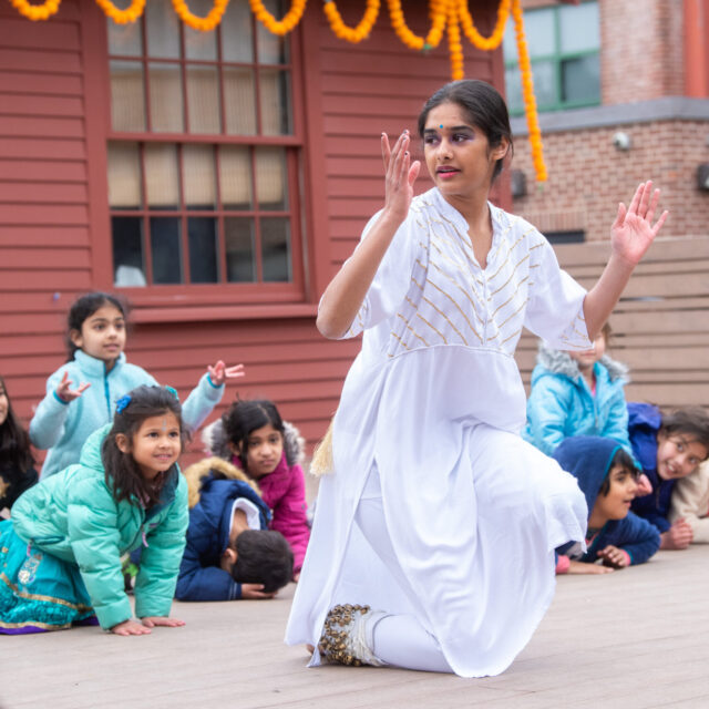 A dancer in a white outfit performs a traditional dance on stage, while a group of children watches attentively in the background.