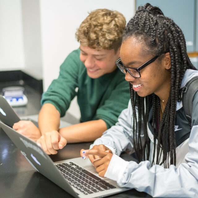 Two students are working together on laptops at a desk, smiling and pointing at their screens.