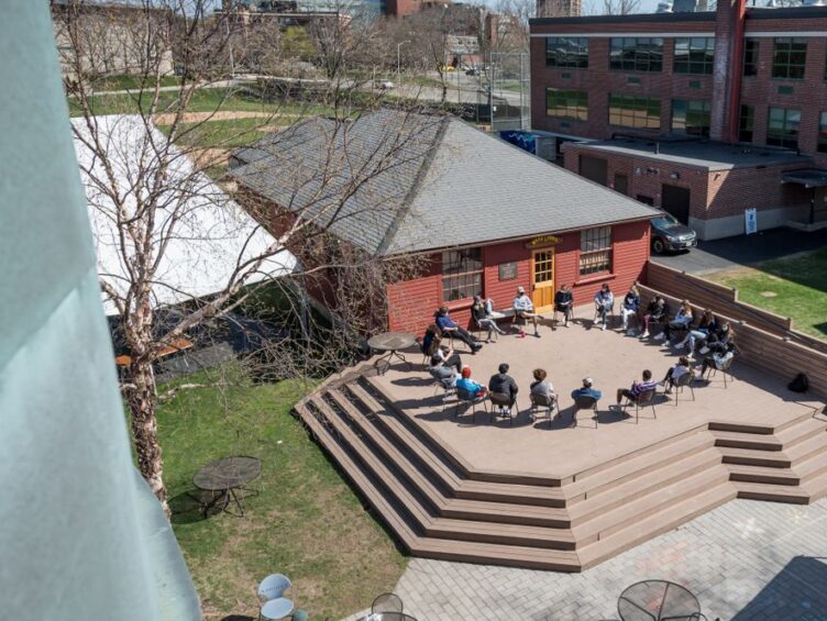 A group of people sit in a circle on an outdoor terrace with steps, next to a small building and green lawn, engaging in a discussion.