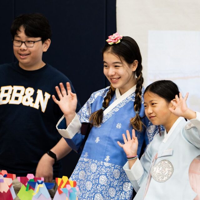 Three children stand together, smiling and waving. Two girls wear traditional attire with braided hair, while the boy stands next to them in a casual outfit.