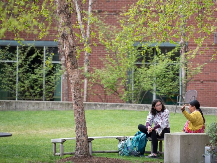 Two people sit on a bench on a grassy campus area, engaging in conversation under trees with a brick building in the background.