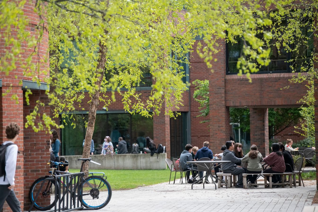 A group of people sit around tables in a shaded outdoor area beside a brick building, while a person walks past a parked bicycle in the foreground.