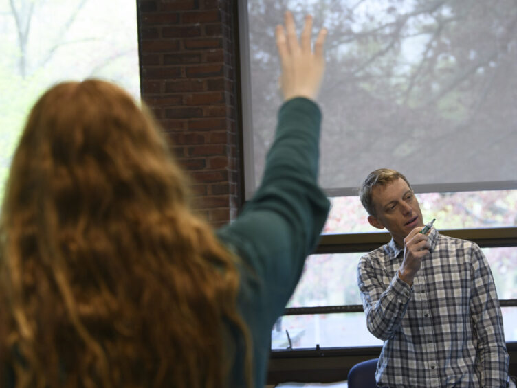 A person with raised hand faces a man holding a marker, who stands in front of a classroom window.