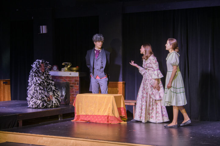 Four people are on a stage performing a scene from a play.