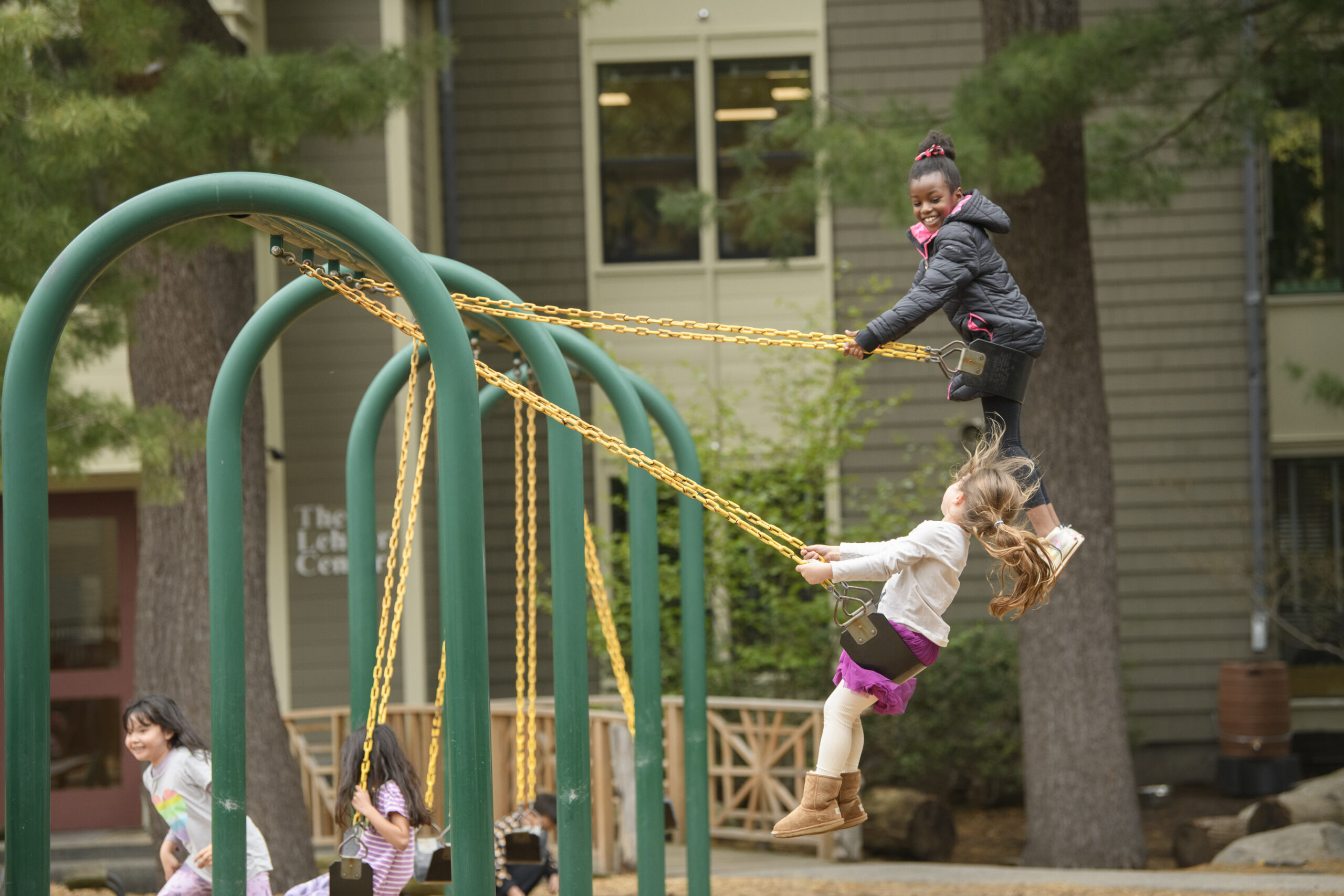 Children playing on swings at a playground. Two girls share a swing, one standing and holding chains while the other is seated.