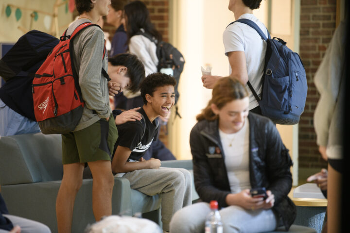 A group of students with backpacks socializing indoors; some are standing while others are seated, and one student is smiling and laughing.