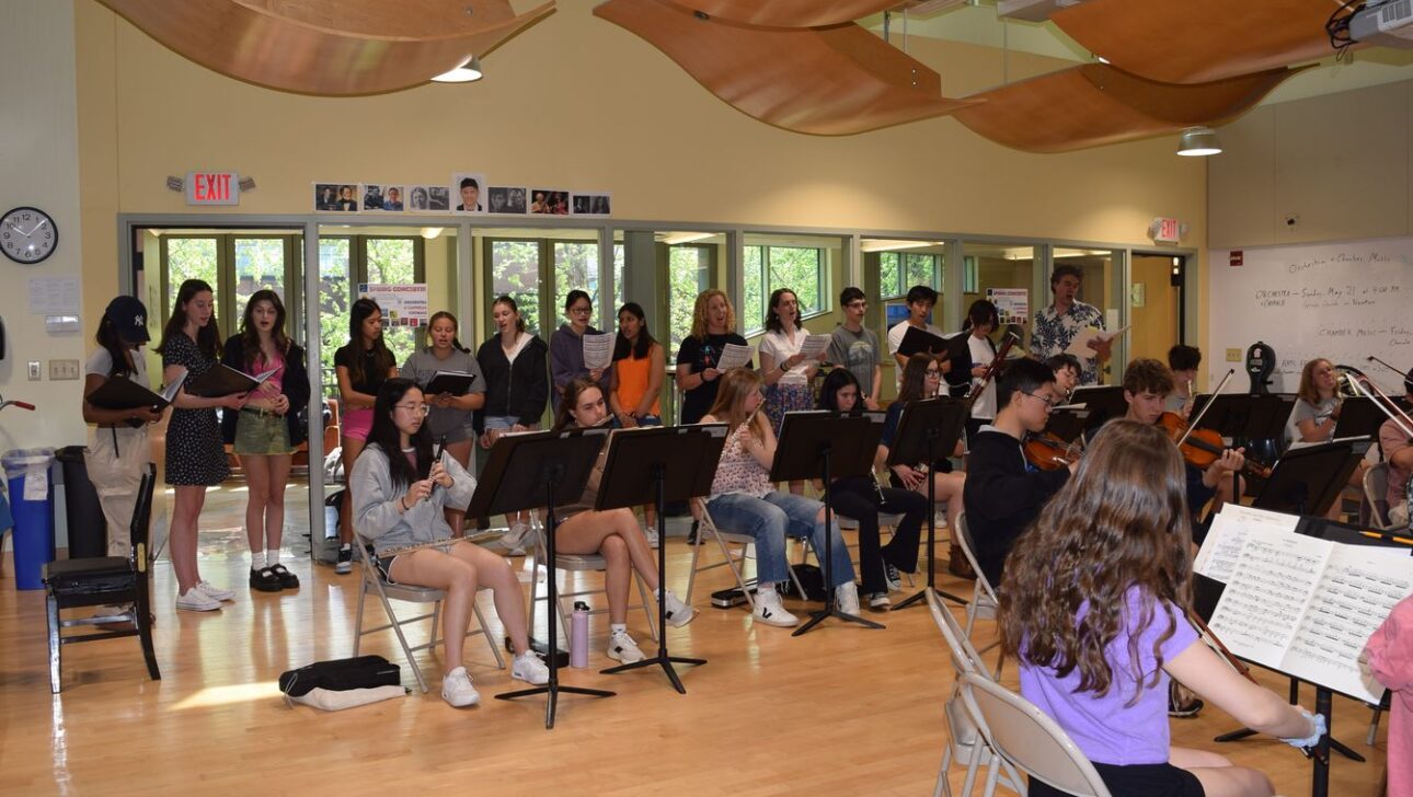A group of students is practicing music in a classroom. Some students are playing instruments while others are singing or observing.