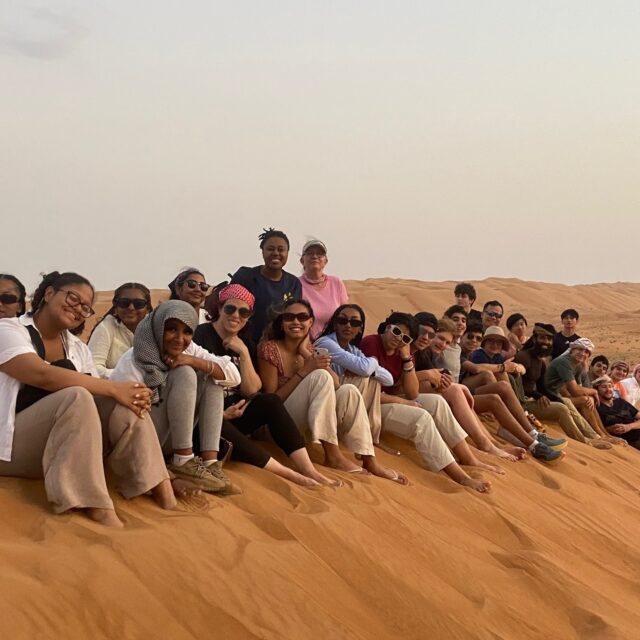 A group of people sitting in a line on a sand dune in a desert, smiling and posing for the camera.