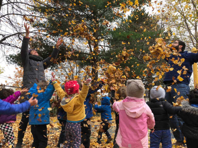 Adults and children, bundled in winter clothes, playfully throw autumn leaves in the air in a park, surrounded by trees with fall foliage.