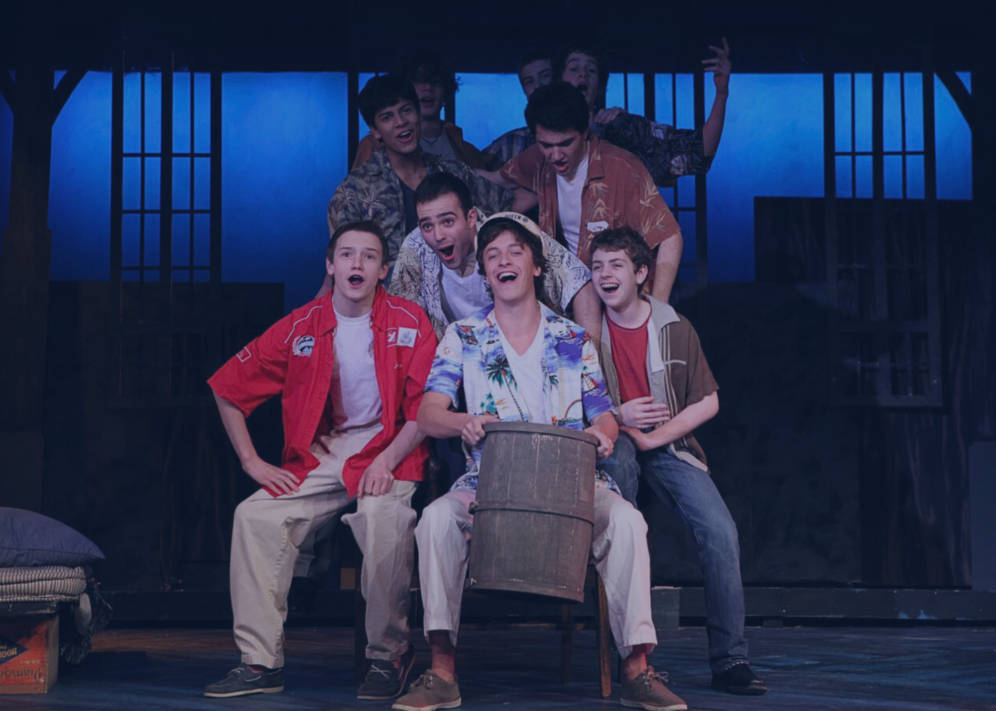 A group of male actors, some standing and some sitting, perform energetically on stage with props and dramatic lighting.
