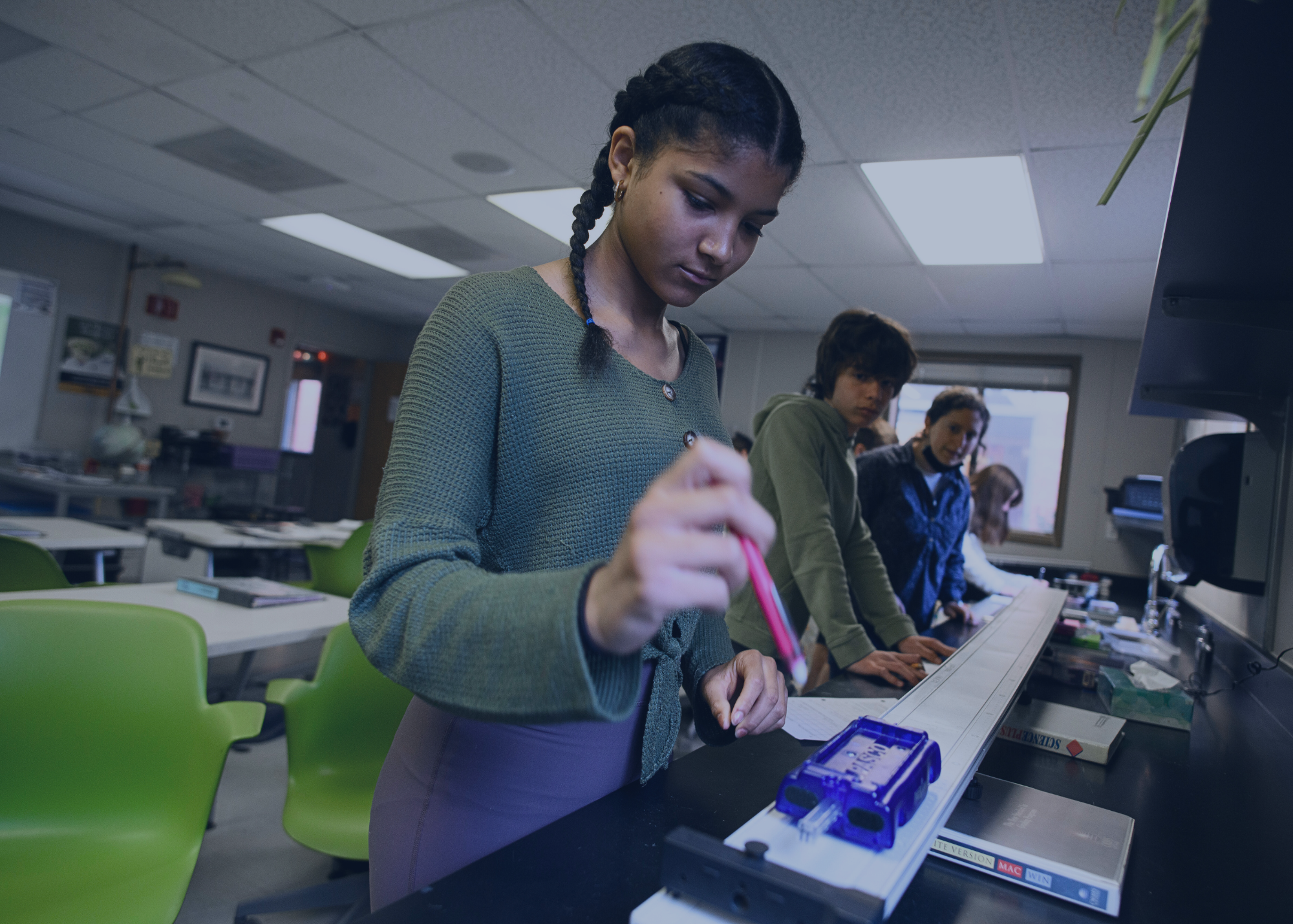 A person with braided hair performs an experiment with a small car on a track in a classroom, while others watch and participate.