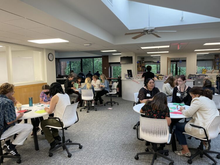 A group of people sits at multiple round tables in a modern, brightly-lit room, engaging in discussions and activities.