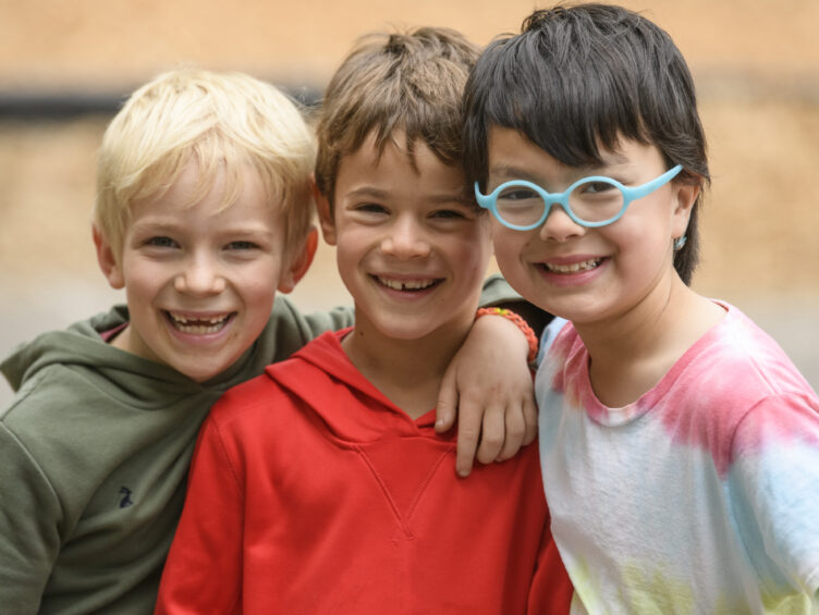 Three smiling children with arms around each other. One wears a green hoodie, another a red hoodie, and the third a colorful tie-dye shirt and blue glasses.
